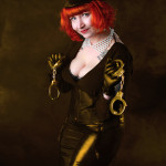 Madam Helle wearing an officer's cap, latex skirt, leather shirt and leather gloves. She is smiling suggestively into the camera and holding two sets of metal handcuffs.
