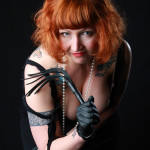 Madam Helle leaning into the camera. She has a short red bob with a full fringe and is wearing red lipstick, black lingerie and black leather gloves. She is holding a small leather flogger.