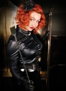 Madam Helle with curly red hair wearing a tight leather shirt and leather gloves. She is looking suggestively into the camera and in one hand holds a black riding crop.