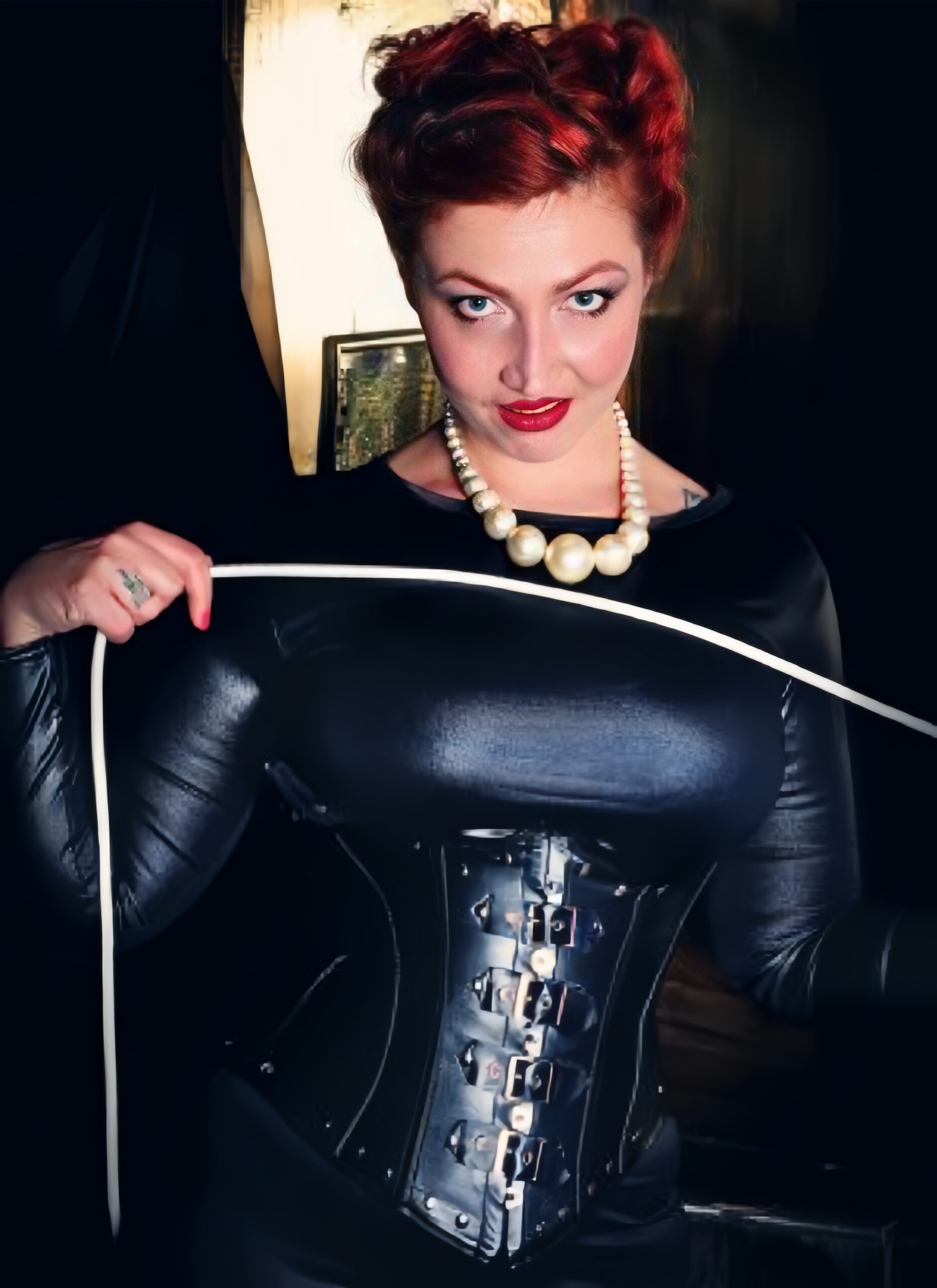 Front-facing photo of Madam Helle, wearing a tight black dress, black corset, and a string of pearls. She is looking into the camera and holding a white rope.