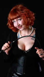 Front-facing photo of Madam Helle, smiling and looking into the camera. She is wearing a black blouse that shows her cleavage and is bending a riding crop between her hands.